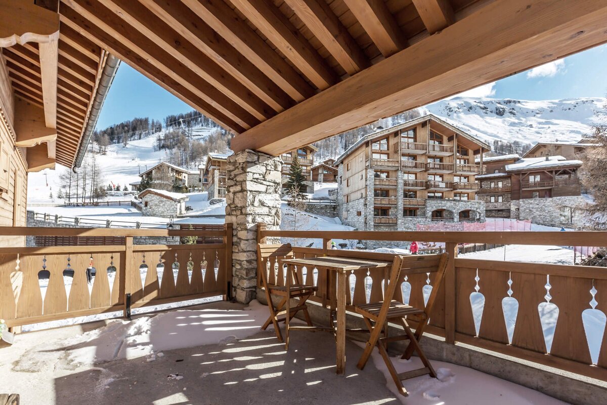 A winter scene from a balcony overlooking a snowy mountain village of chalets and ski slopes under a blue sky.