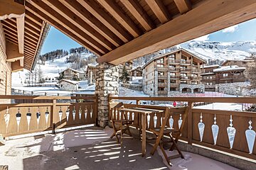 A winter scene from a balcony overlooking a snowy mountain village of chalets and ski slopes under a blue sky.