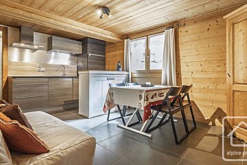 A cozy, modern chalet-style room with wooden walls/ceiling, a kitchenette, dining table, and sofa. A winter scene is visible through the window.