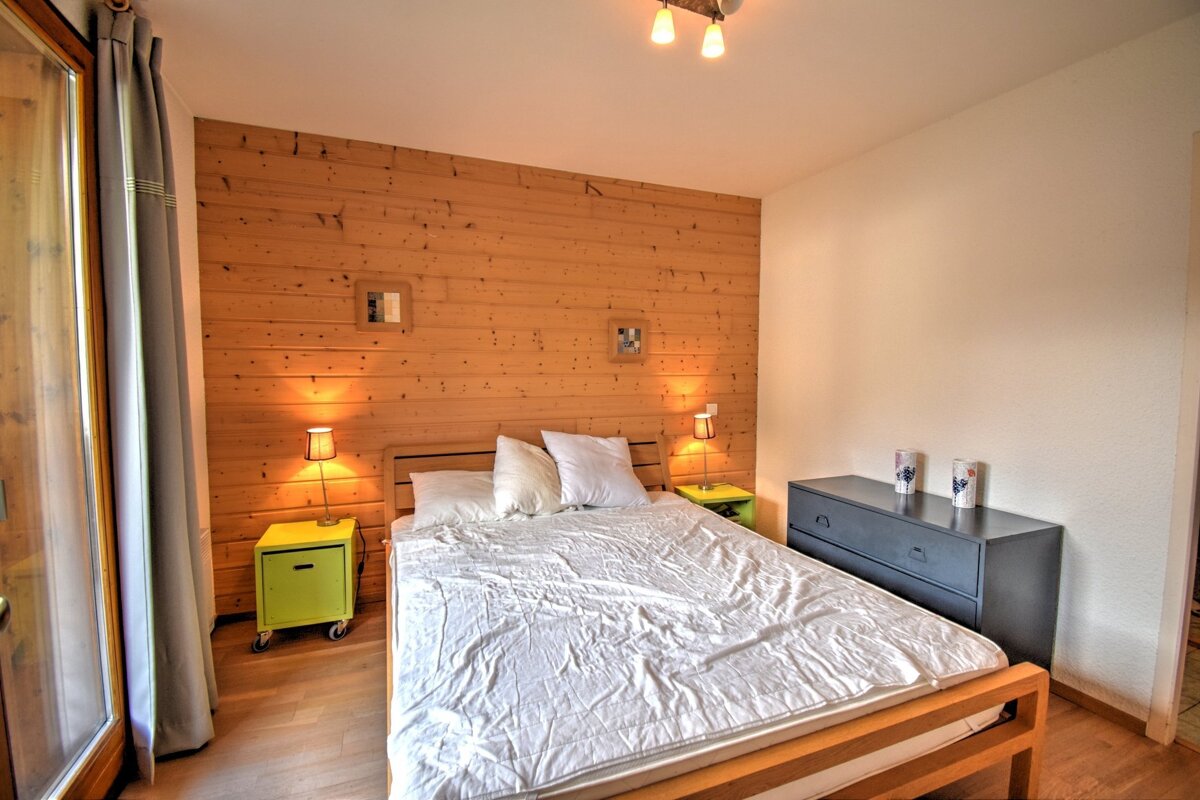 A cozy bedroom with a wooden accent wall, a double bed with white linens, bright nightstands, and a dark dresser. Warm lighting.