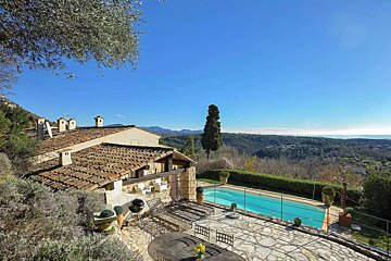 A Mediterranean villa with a tiled roof, stone patio, and swimming pool, overlooking scenic hills and a clear blue sky.