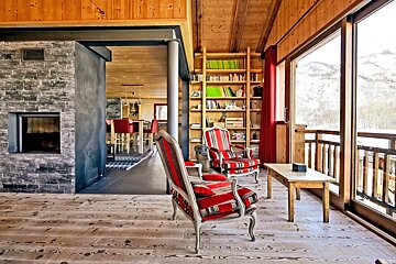 Cozy alpine chalet living room featuring red-striped armchairs, a stone fireplace, and large windows overlooking snowy mountains.