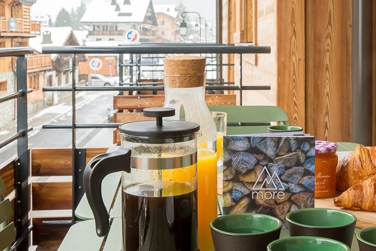 Breakfast on an alpine balcony featuring a French press, orange juice, and croissants, overlooking a snowy mountain village.