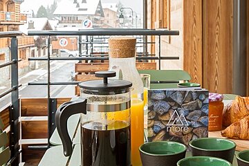 Breakfast on an alpine balcony featuring a French press, orange juice, and croissants, overlooking a snowy mountain village.