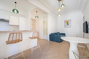 A bright, modern open-plan apartment with white walls, a blue sofa, light wood floors, and a kitchen island with barstools.