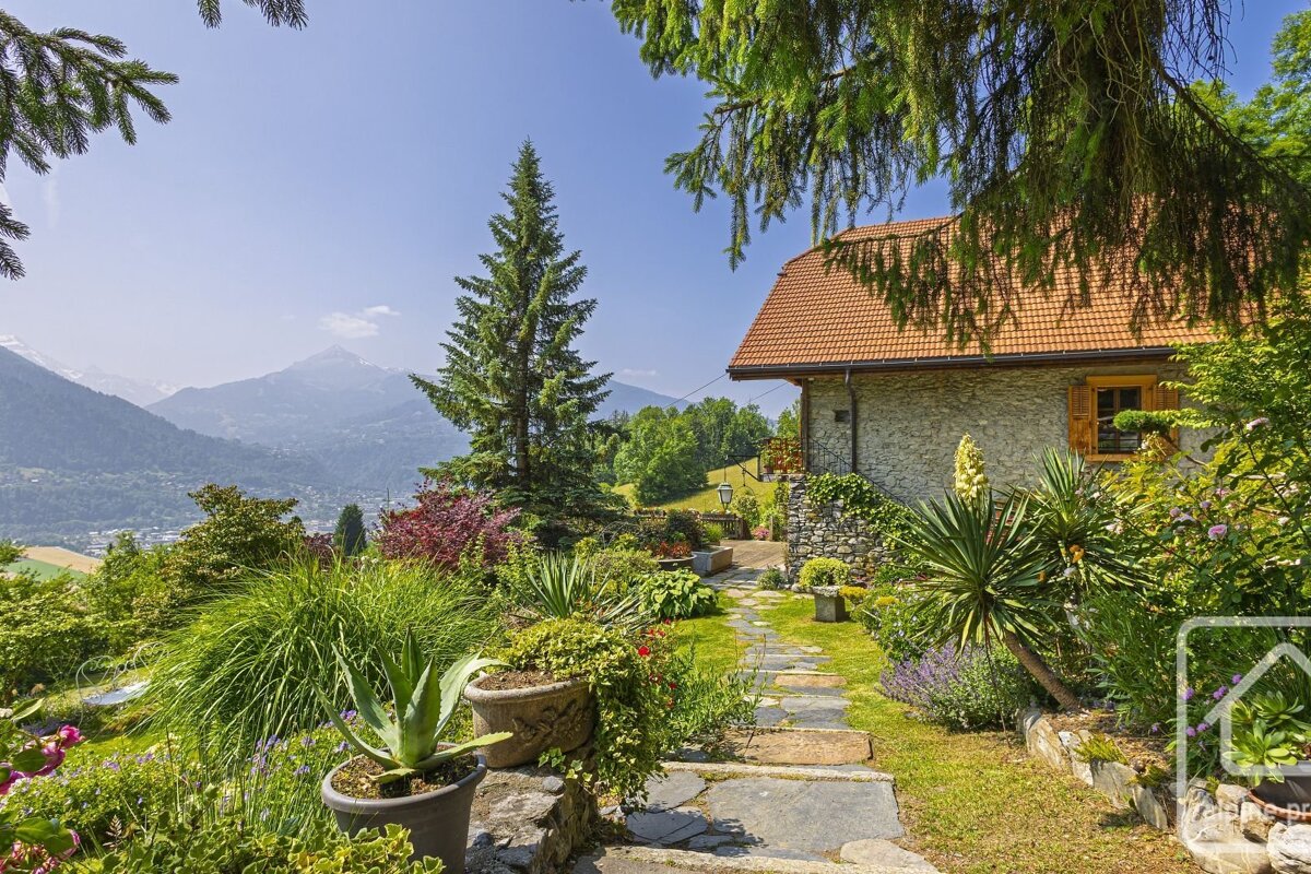 A stone house with a red roof sits in a vibrant garden with a path, overlooking a scenic mountain valley under a clear blue sky.