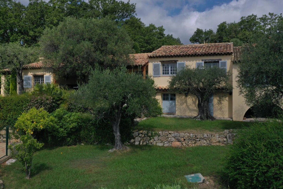 A charming Mediterranean-style house with blue shutters and a tiled roof, surrounded by olive trees and lush green lawns.