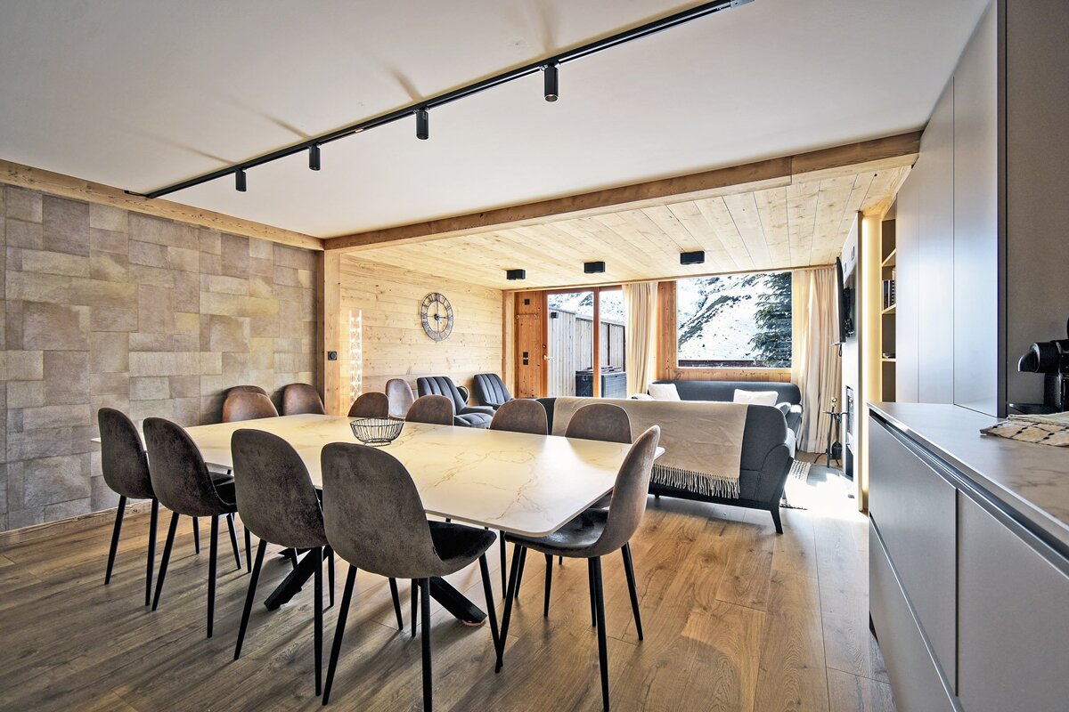 Modern chalet interior with a marble dining table, grey chairs, wood paneling, and a living area with snowy mountain views.