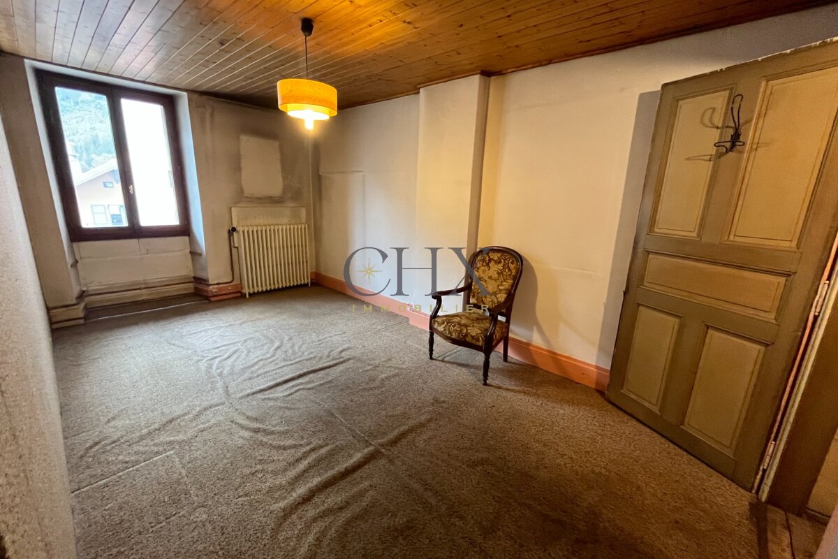 An empty room with a wooden ceiling, brown carpet, a single vintage armchair, a yellow hanging lamp, a window, and a wooden door.