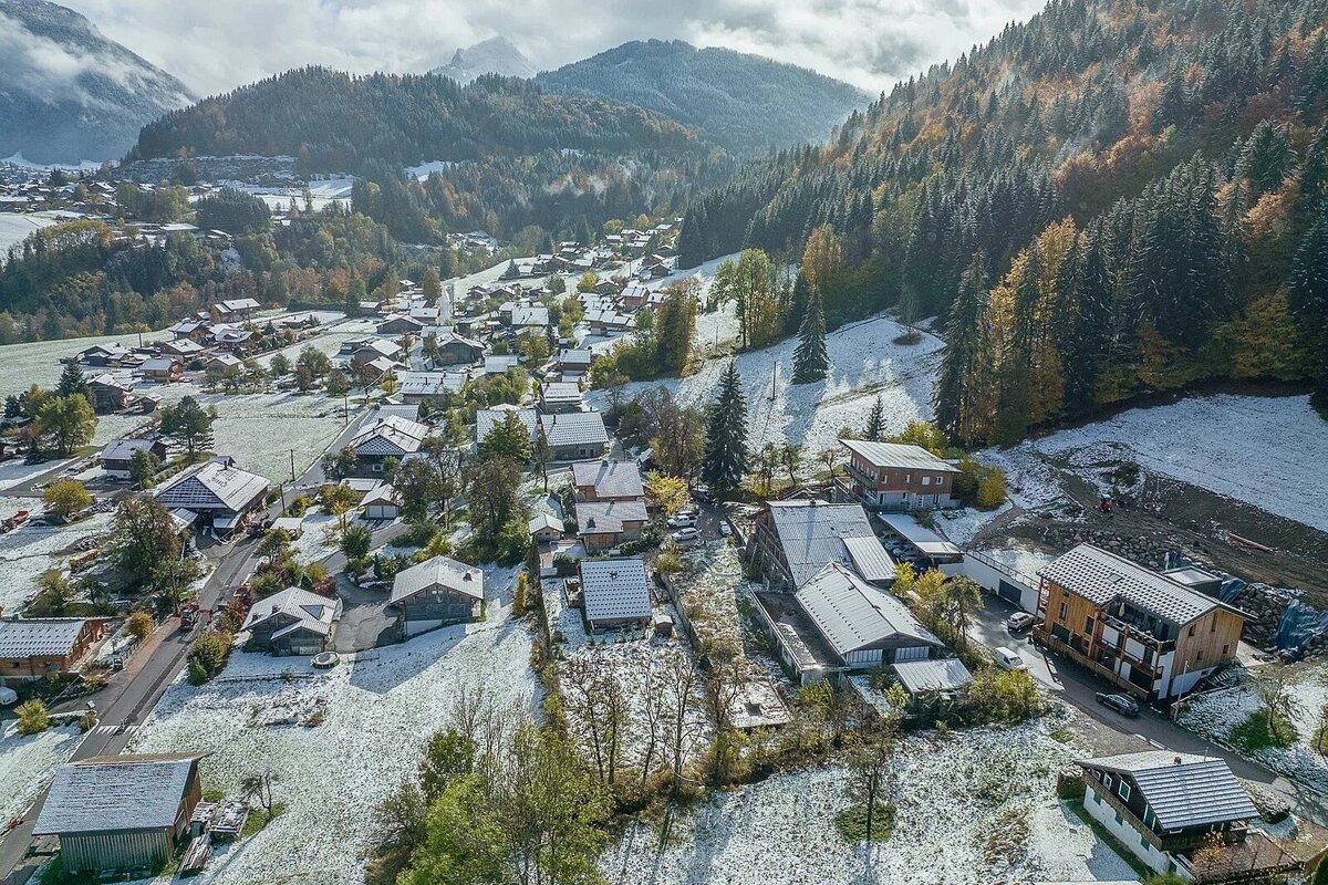 Aerial view of a snowy mountain village nestled among evergreen and autumn-colored trees, with cloudy skies overhead.