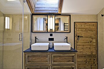 A rustic modern bathroom featuring a double vanity with white sinks on a wooden cabinet, beige tiled walls, a glass shower, and a wooden door.