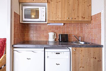 A compact kitchen featuring wood cabinets, terracotta tiles, microwave, hob, sink, kettle, toaster, fridge, and dishwasher.