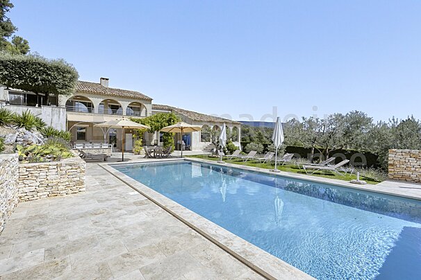 A large swimming pool is surrounded by chairs and umbrellas in front of a large house