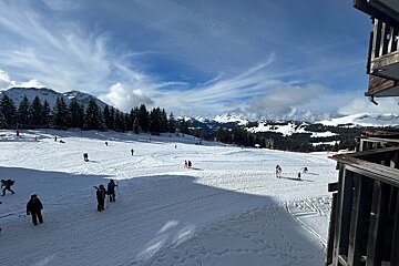A snowy ski resort with skiers and snowboarders on the slopes under a blue sky, framed by a wooden balcony.