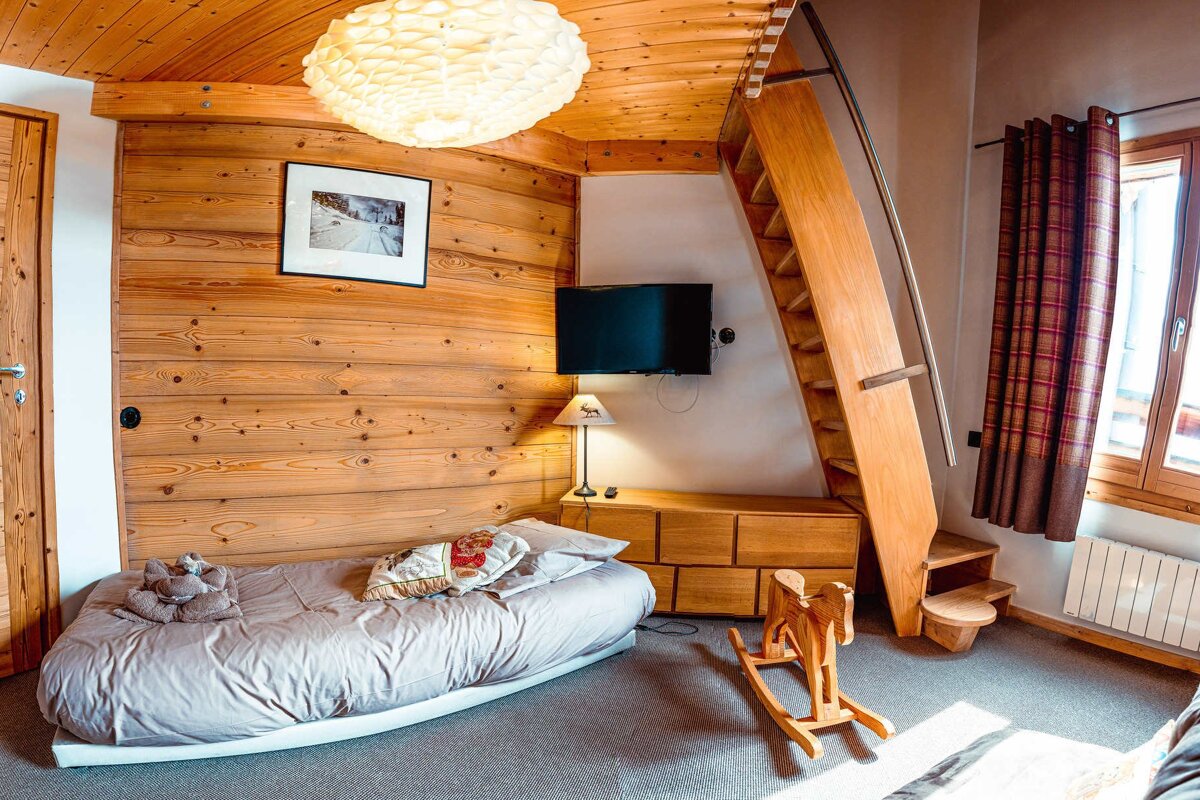 Cozy, wood-paneled room with a single bed, wall-mounted TV, unique wooden ladder/staircase, and a rocking horse on a gray carpet.