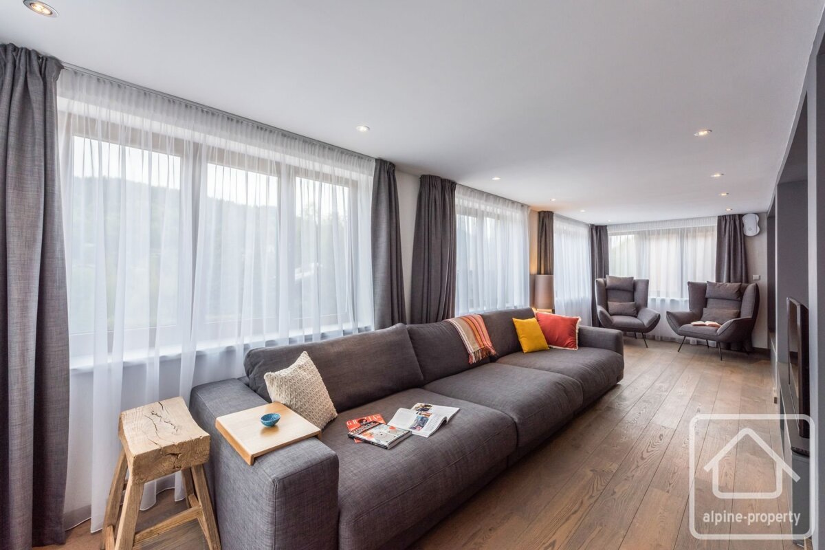 A modern living room with a long gray sectional sofa, hardwood floor, and large windows with sheer and gray curtains. Two armchairs are visible.