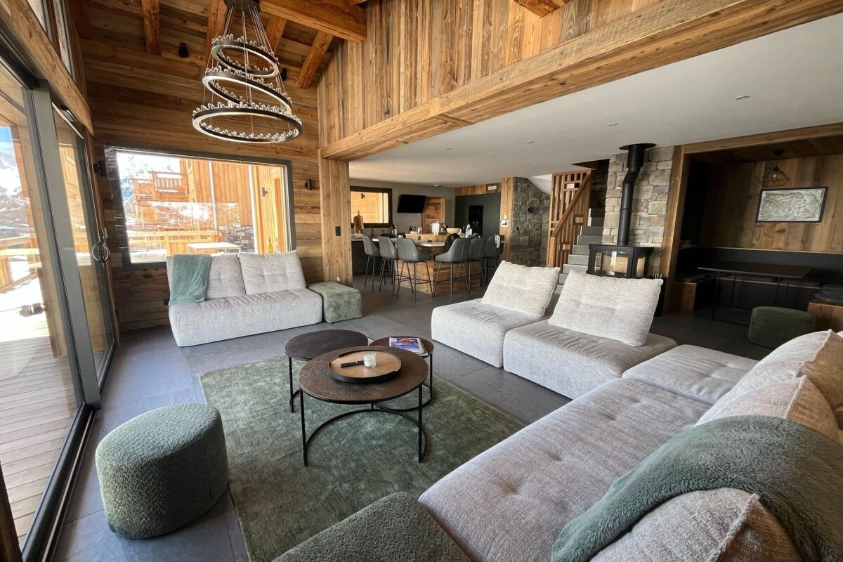Cozy wooden chalet interior with plush grey sofas, a green rug, nesting coffee tables, a modern chandelier, and large scenic windows.