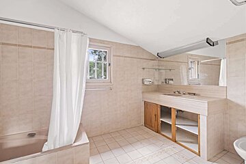 A tiled bathroom features a bathtub with a white shower curtain, a vanity with a sink, a large mirror, and a window.