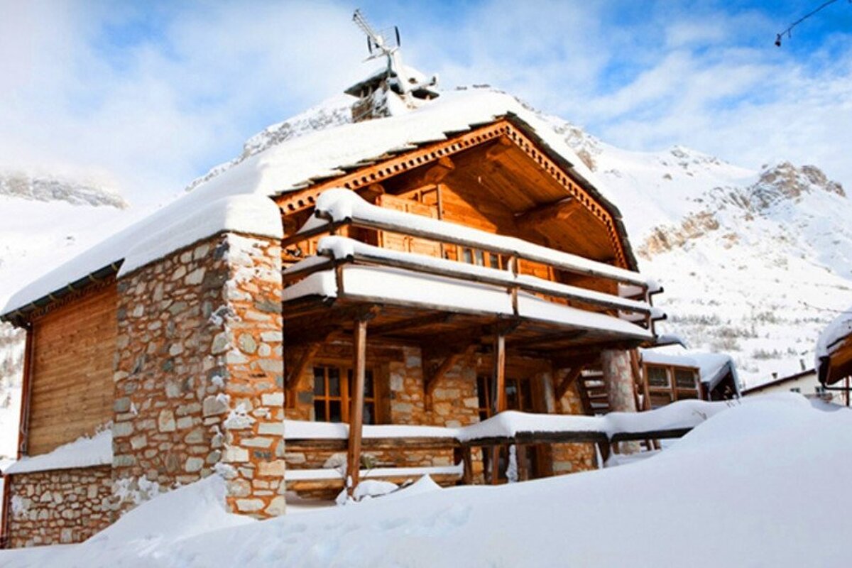 A rustic wooden and stone cabin covered in thick snow, nestled in a snowy mountain landscape under a blue sky.