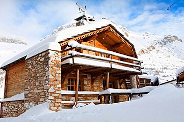A rustic wooden and stone cabin covered in thick snow, nestled in a snowy mountain landscape under a blue sky.