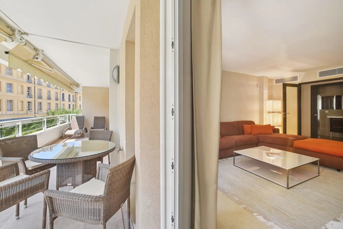 A modern balcony with outdoor seating and city views, connected to an indoor living room featuring a brown sectional sofa and coffee table.