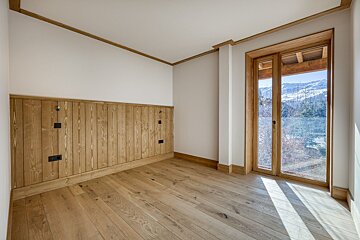 Empty chalet room with light wood floors, wood paneling, and a glass door looking out to a snowy mountain view.