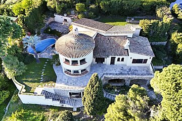 Aerial view of a grand villa with a round tower, terracotta roofs, and a blue swimming pool, nestled among lush green trees and terraced gardens.