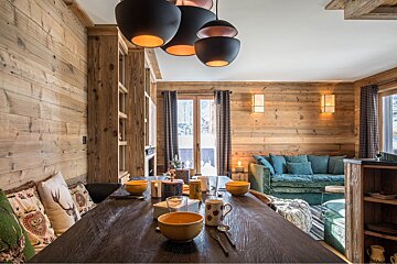 A cozy, rustic wood-paneled chalet interior with a dining table set, a teal sofa, and black pendant lights, looking out to a snowy landscape.