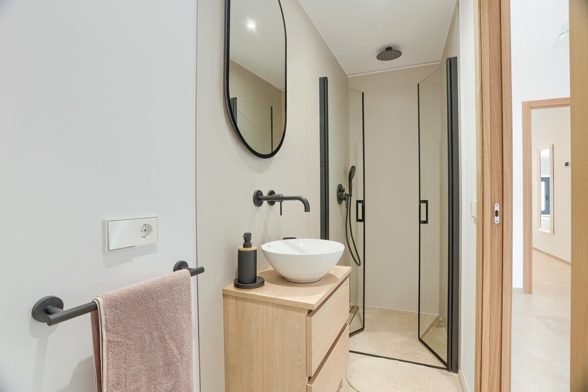 A modern bathroom featuring light walls, black fixtures, a wooden vanity with a white basin, an oval mirror, and a sleek glass shower.