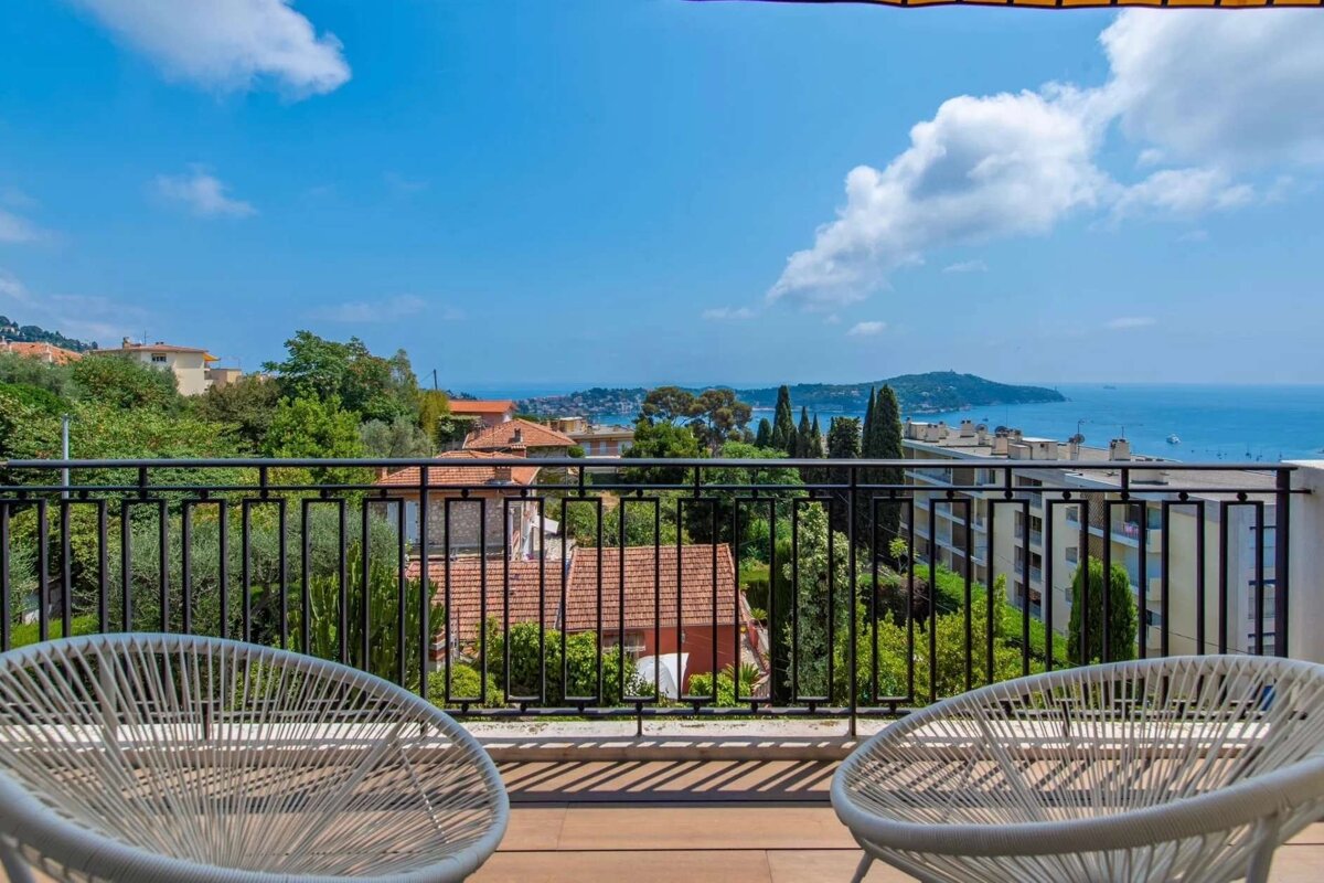 A sunny balcony with two white chairs offers a stunning panoramic view of a coastal town, lush green landscape, and the blue Mediterranean Sea.