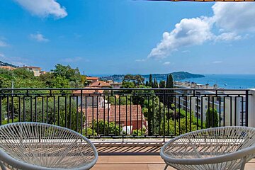 A sunny balcony with two white chairs offers a stunning panoramic view of a coastal town, lush green landscape, and the blue Mediterranean Sea.