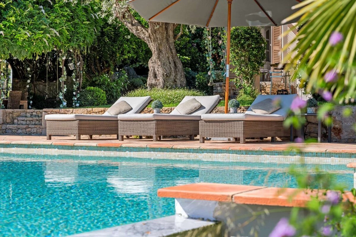 A tranquil outdoor pool with several wicker lounge chairs under a large umbrella, surrounded by lush green foliage and trees. The clear blue water sparkles.
