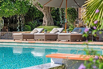 A tranquil outdoor pool with several wicker lounge chairs under a large umbrella, surrounded by lush green foliage and trees. The clear blue water sparkles.