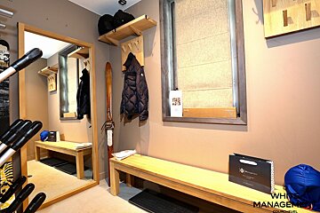 A cozy ski lodge entryway with wooden benches, a mirror, coat rack, ski decor, and window, all in warm neutral tones. Ski poles and bags visible.