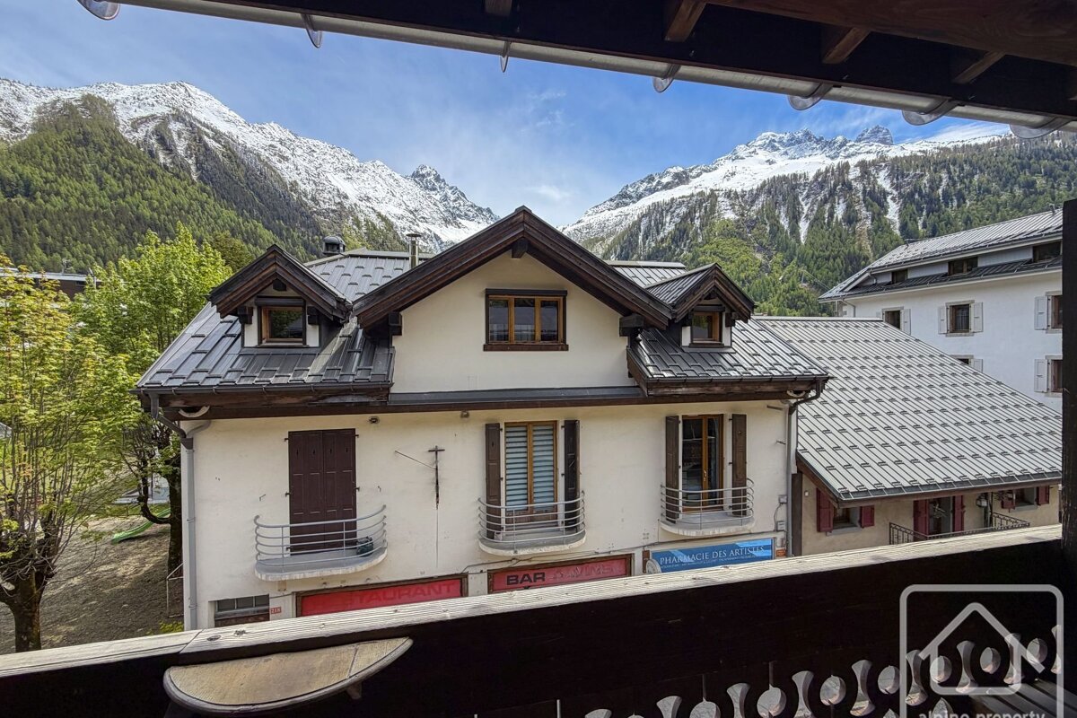 Alpine scene from above: a building with shops, green trees, and majestic snow-capped mountains under a blue sky.