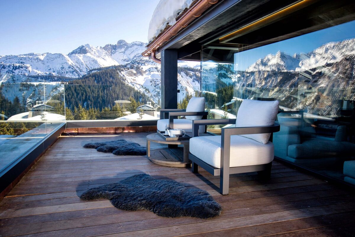 Modern balcony with outdoor seating and sheepskin rugs, offering a panoramic view of snowy mountains, pine forests, and a clear blue sky.