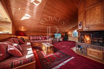Cozy wood-paneled attic room with a red carpet, striped sofas, a burning fireplace, TV, and a mountain view from the window.