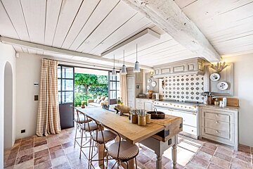 Charming rustic kitchen with a long wooden island, exposed beams, tiled floor, and a view of a lush garden through open French doors.