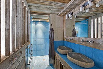 Rustic bathroom with wood paneling, vibrant blue tiles, unique stone sinks, and a glass-enclosed shower.