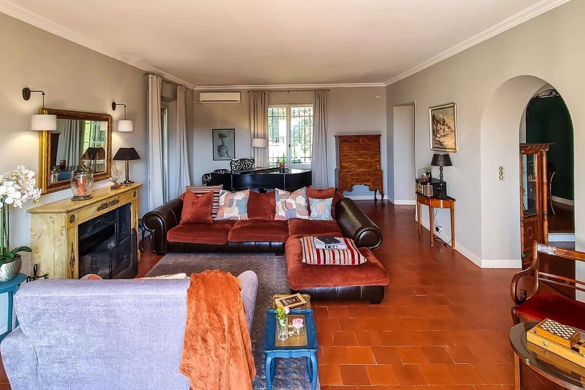 A spacious living room with a red sectional sofa, terracotta floor, fireplace, grand piano, and classic wooden furniture, creating an inviting ambiance.