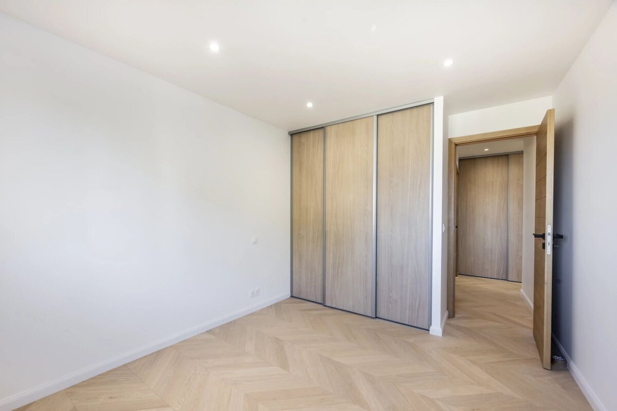 An empty, bright room with white walls, chevron wood floors, and light wood built-in sliding wardrobes beside an open doorway.