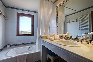 A tiled bathroom features a tub by a window with a mountain view, a vanity with a sink, mirror, and towels. Blue and white decor.