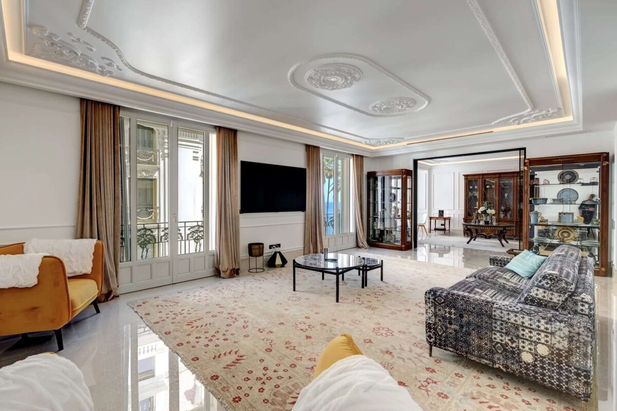 A luxurious living room featuring ornate ceiling moldings, balcony access, a large TV, patterned rug, elegant furniture, and a distant sea view.
