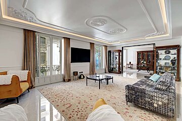 A luxurious living room featuring ornate ceiling moldings, balcony access, a large TV, patterned rug, elegant furniture, and a distant sea view.