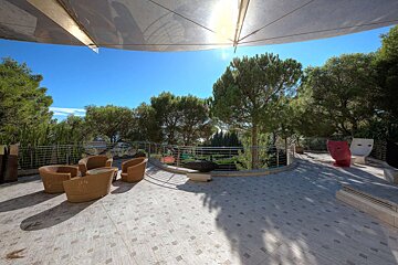A sunlit outdoor patio with wicker furniture under a large canopy. Lush trees surround a metal railing, with decorative face planters visible. Blue sky.