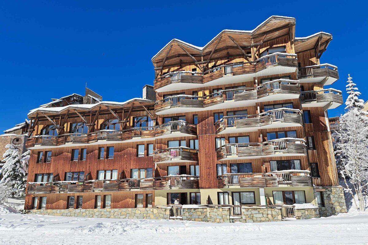 A snow-covered building, likely a hotel, with icicles and parked cars in front, set against snowy mountains and pine forests in winter.