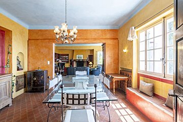 A rustic, warm-toned dining room features orange and yellow walls, a glass-top dining table, and a view into a living room with a fireplace.
