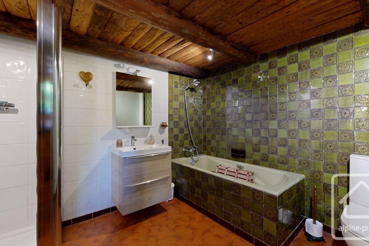 A rustic bathroom featuring exposed wooden ceiling beams, white and green patterned tiles, a bathtub, sink, and hexagonal terracotta floor.