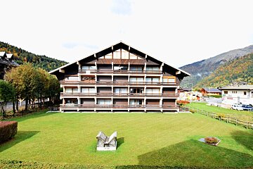 A large chalet-style building with multiple balconies, a green lawn, and autumn mountains in the background under a bright sky.