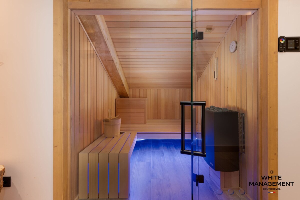 A modern wood-paneled sauna with a glass door, blue LED floor lighting, a heater, and a wooden bench with a water bucket.